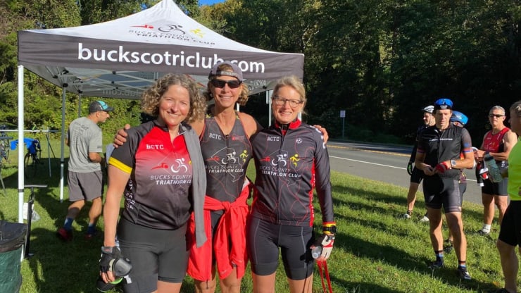 Three members of Bucks County Tri Club pose for a photo in front of a tent that has buckscotriclub.com printed on the side.